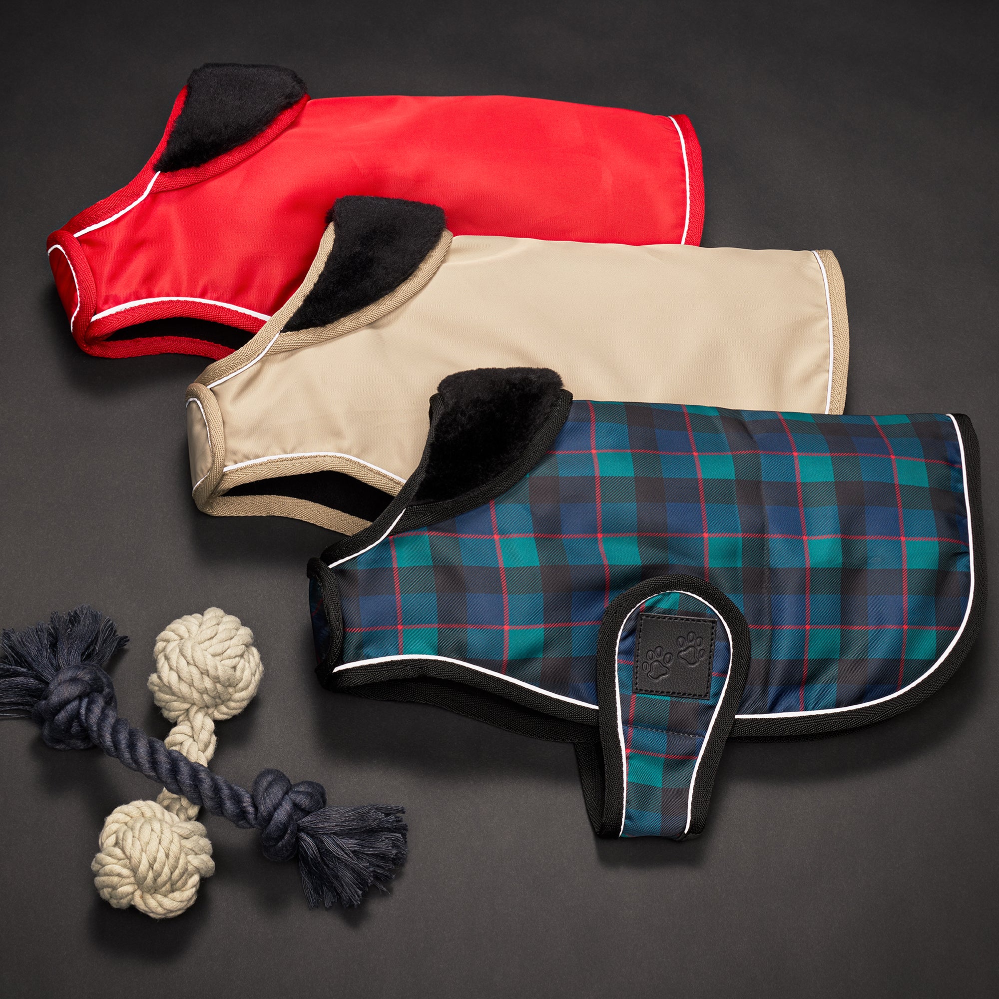 Three dog coats in red, beige, and plaid pattern with two rope toys on a dark background